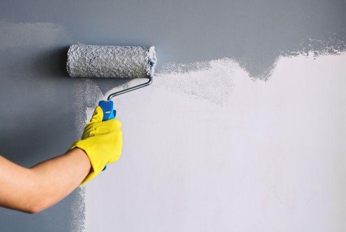 Une main avec un gant jaune peint un mur en gris avec un rouleau à peinture