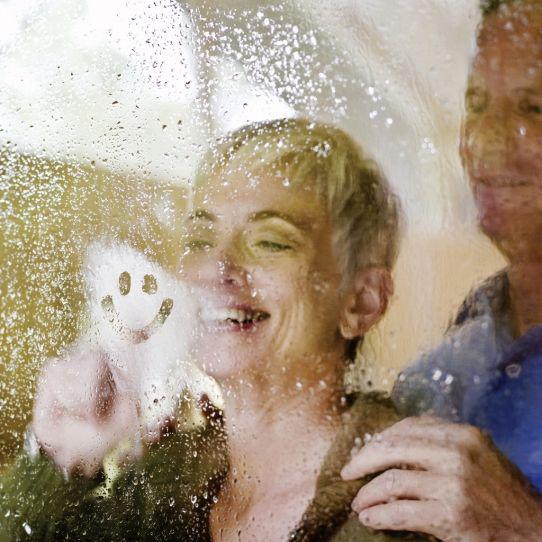 Un couple souriant regarde à travers une fenêtre humide et embuée tandis que la femme dessine un smiley à l'intérieur